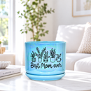 A light blue ceramic plant pot with a matching saucer, decorated with black line drawings of potted plants and the text 'Best Mom ever', sitting on a light-colored table in a bright, softly blurred living room setting.