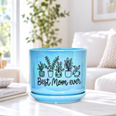 A light blue ceramic plant pot with a matching saucer, decorated with black line drawings of potted plants and the text 'Best Mom ever', sitting on a light-colored table in a bright, softly blurred living room setting.