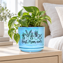 A light blue ceramic plant pot with a matching saucer, decorated with black line drawings of potted plants and the text 'Best Mom ever', sitting on a light-colored table in a bright, softly blurred living room setting.