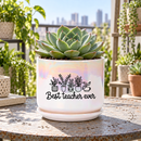 A white ceramic planter pot with a matching saucer sits on a dark kitchen countertop. The pot has an iridescent sheen and is printed with a black graphic of potted plants and the text 'Best teacher ever'.