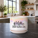 A white ceramic planter pot with a matching saucer sits on a dark kitchen countertop. The pot has an iridescent sheen and is printed with a black graphic of potted plants and the text 'Best teacher ever'.
