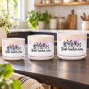 A white ceramic planter pot with a matching saucer sits on a dark kitchen countertop. The pot has an iridescent sheen and is printed with a black graphic of potted plants and the text 'Best teacher ever'.