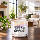 A white ceramic planter pot with a saucer, featuring a black graphic of potted plants and the word 'GRANDMA' in script, set on a wooden table in a bright, modern living room.