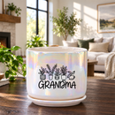 A white ceramic planter pot with a saucer, featuring a black graphic of potted plants and the word 'GRANDMA' in script, set on a wooden table in a bright, modern living room.