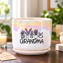 A white ceramic planter pot with a saucer, featuring a black graphic of potted plants and the word 'GRANDMA' in script, set on a wooden table in a bright, modern living room.