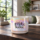 An iridescent ceramic planter pot with a matching saucer sits on a dark wooden table in a cozy living room setting. The pot features a black graphic of potted plants and the word 'MAMA' printed on the front.