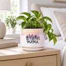 An iridescent ceramic planter pot with a matching saucer sits on a dark wooden table in a cozy living room setting. The pot features a black graphic of potted plants and the word 'MAMA' printed on the front.
