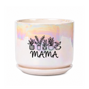 An iridescent ceramic planter pot with a matching saucer sits on a dark wooden table in a cozy living room setting. The pot features a black graphic of potted plants and the word 'MAMA' printed on the front.