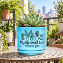 A light blue ceramic planter with a matching saucer, featuring a graphic of potted plants and the text 'My life would succ without you', displayed on a wooden table in a bright, sunlit room next to a vase of white flowers.