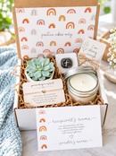 A curated gift box set with a small succulent, a glass jar candle, cards with positive quotes, and a branded box lid printed with repeating rainbow motifs and the phrase 'positive vibes only'. The box is styled on a light surface with a blue knit blanket nearby.