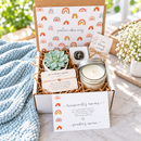 A curated gift box set with a small succulent, a glass jar candle, cards with positive quotes, and a branded box lid printed with repeating rainbow motifs and the phrase 'positive vibes only'. The box is styled on a light surface with a blue knit blanket nearby.