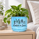 A light blue ceramic planter pot with a matching saucer, featuring a black line-art design of potted plants and the text 'Rooted in Love'. The background shows a softly blurred bookshelf with books and small plants.