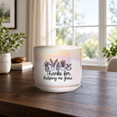 A white ceramic planter pot with a subtle iridescent sheen, featuring a black graphic of potted plants and the text 'Thanks for helping me grow' on the front. The pot sits on a dark wooden table in a bright, modern living room setting with a window, plants, and curtains in the background.