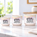 A white ceramic planter pot with a subtle iridescent sheen, featuring a black graphic of potted plants and the text 'Thanks for helping me grow' on the front. The pot sits on a dark wooden table in a bright, modern living room setting with a window, plants, and curtains in the background.