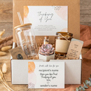 A gift box set containing a jar candle with a handwritten label, a small succulent in a white pot, and a matchstick tube, all nestled in brown crinkle paper inside an open white cardboard box.