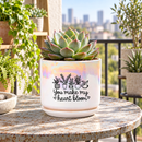 A ceramic planter pot with a matching saucer sits on a dark countertop in a bright, modern living room. The pot is white with a holographic iridescent finish on the upper half, decorated with black line drawings of potted plants and the text 'You make my heart bloom'.