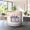 A ceramic planter pot with a matching saucer sits on a dark countertop in a bright, modern living room. The pot is white with a holographic iridescent finish on the upper half, decorated with black line drawings of potted plants and the text 'You make my heart bloom'.