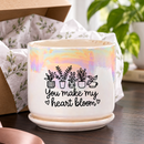 A ceramic planter pot with a matching saucer sits on a dark countertop in a bright, modern living room. The pot is white with a holographic iridescent finish on the upper half, decorated with black line drawings of potted plants and the text 'You make my heart bloom'.
