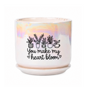 A ceramic planter pot with a matching saucer sits on a dark countertop in a bright, modern living room. The pot is white with a holographic iridescent finish on the upper half, decorated with black line drawings of potted plants and the text 'You make my heart bloom'.