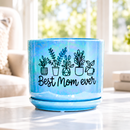 A light blue ceramic plant pot with a matching saucer, decorated with black line drawings of potted plants and the text 'Best Mom ever', sitting on a light-colored table in a bright, softly blurred living room setting.