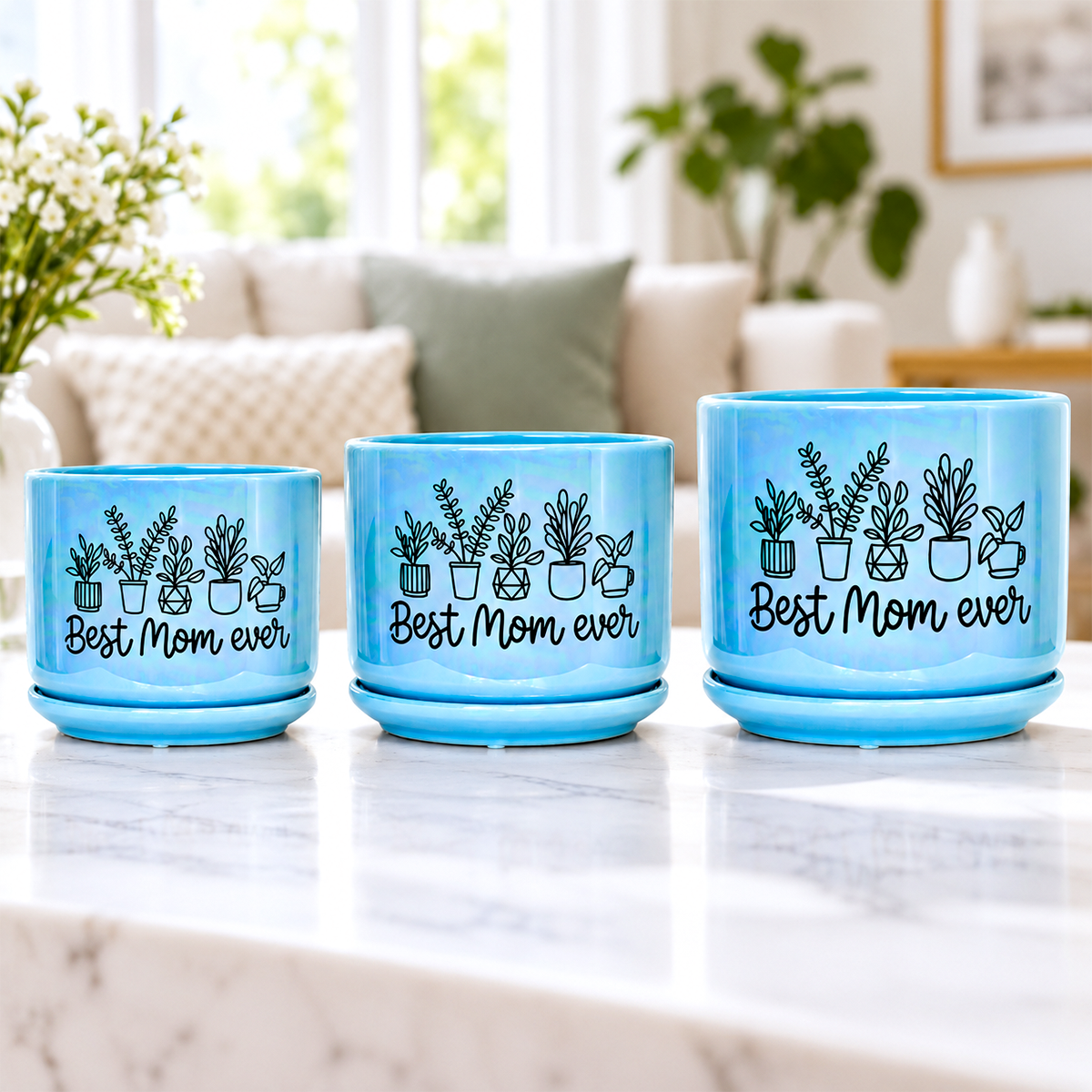 Three light blue ceramic planters with saucers on a white marble countertop, featuring black illustrations of potted plants and the text 'Best Mom ever' on the front, set against a blurred living room background.