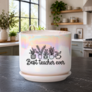 A white ceramic planter pot with a matching saucer sits on a dark kitchen countertop. The pot has an iridescent sheen and is printed with a black graphic of potted plants and the text 'Best teacher ever'.