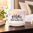 A white ceramic planter pot with a matching saucer sits on a dark kitchen countertop. The pot has an iridescent sheen and is printed with a black graphic of potted plants and the text 'Best teacher ever'.