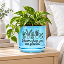 A light blue ceramic planter pot with a matching saucer, featuring a black line art design of potted plants and the text 'Bloom where you are planted,' set on a white table in a bright, modern living room.