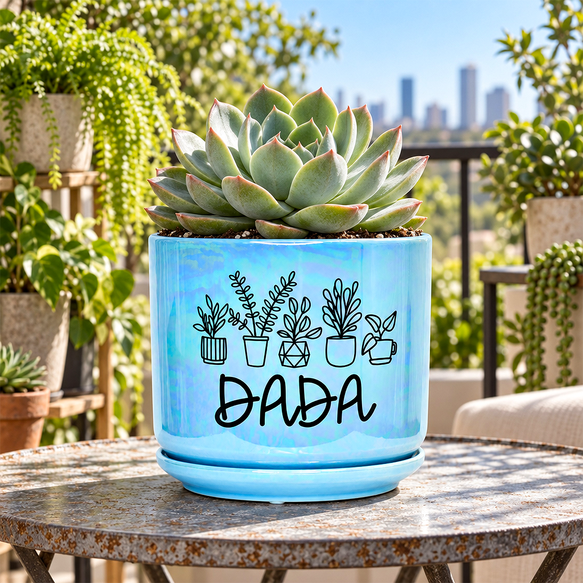 A light blue ceramic planter with a matching saucer, featuring a black line-art design of potted plants and the word 'DADA', holding a green succulent. The planter is displayed on a rustic metal table in a lush garden setting with a city skyline in the background.