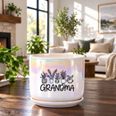 A white ceramic planter pot with a saucer, featuring a black graphic of potted plants and the word 'GRANDMA' in script, set on a wooden table in a bright, modern living room.