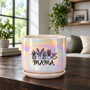 An iridescent ceramic planter pot with a matching saucer sits on a dark wooden table in a cozy living room setting. The pot features a black graphic of potted plants and the word 'MAMA' printed on the front.
