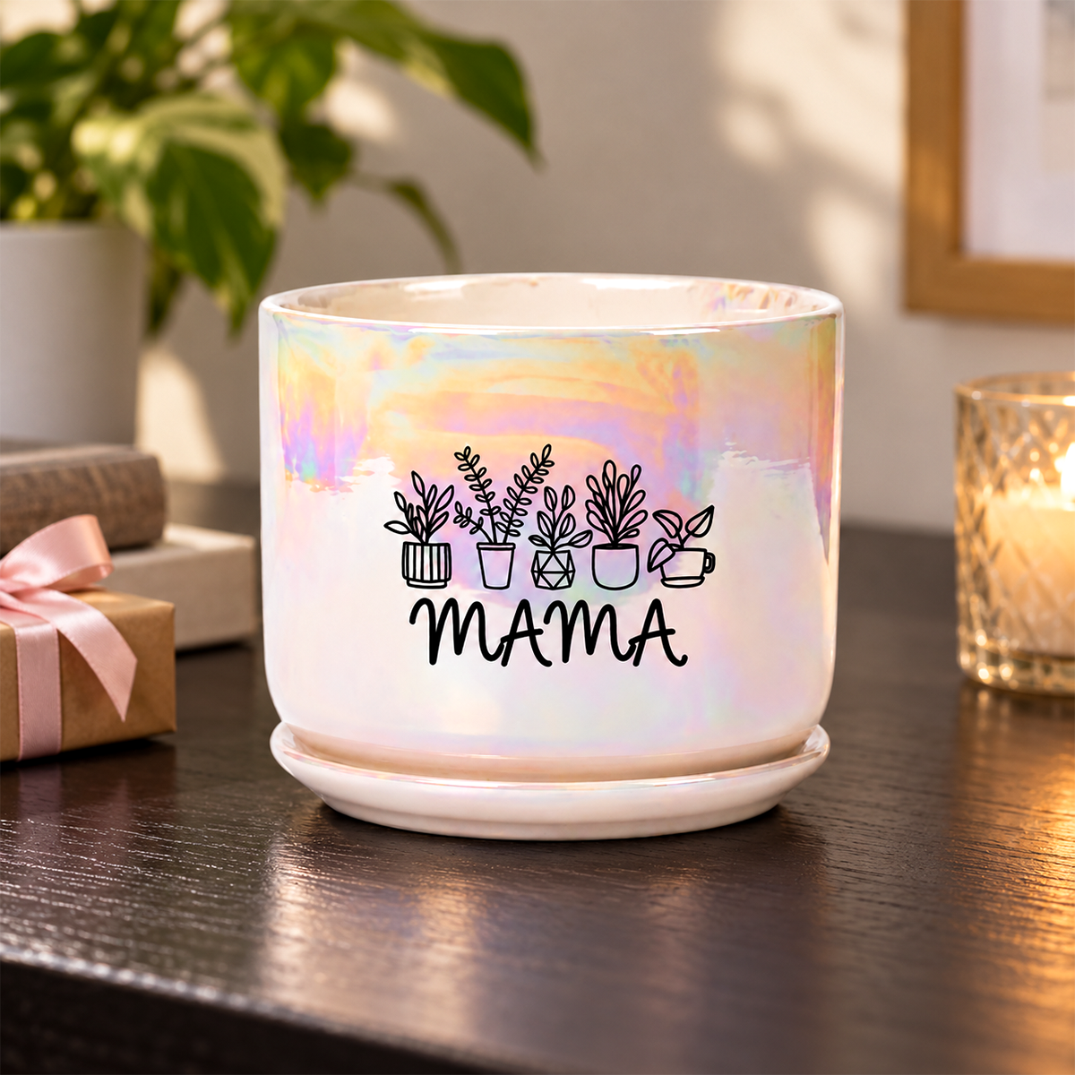 A white ceramic plant pot with a matching saucer, featuring an iridescent pastel rainbow sheen and a black graphic of potted plants above the word 'MAMA'. The pot is set on a dark wooden table in a cozy indoor setting with a lit candle, a gift box, and a houseplant in the background.