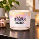 An iridescent ceramic planter pot with a matching saucer sits on a dark wooden table in a cozy living room setting. The pot features a black graphic of potted plants and the word 'MAMA' printed on the front.