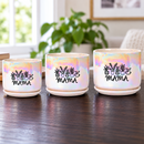 An iridescent ceramic planter pot with a matching saucer sits on a dark wooden table in a cozy living room setting. The pot features a black graphic of potted plants and the word 'MAMA' printed on the front.