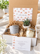 A curated gift box set with a small succulent, a glass jar candle, cards with positive quotes, and a branded box lid printed with repeating rainbow motifs and the phrase 'positive vibes only'. The box is styled on a light surface with a blue knit blanket nearby.