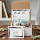 An open gift box filled with self-care and affirmation items, including a black insulated tumbler with 'warrior' printed on it, a candle jar labeled 'breathe', a round sticker reading 'I CAN DO ALL THINGS', a small succulent, and printed cards with motivational text, arranged in brown paper shred on a light surface.