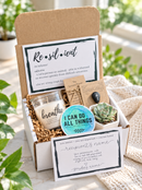An open gift box filled with self-care and affirmation items, including a black insulated tumbler with 'warrior' printed on it, a candle jar labeled 'breathe', a round sticker reading 'I CAN DO ALL THINGS', a small succulent, and printed cards with motivational text, arranged in brown paper shred on a light surface.