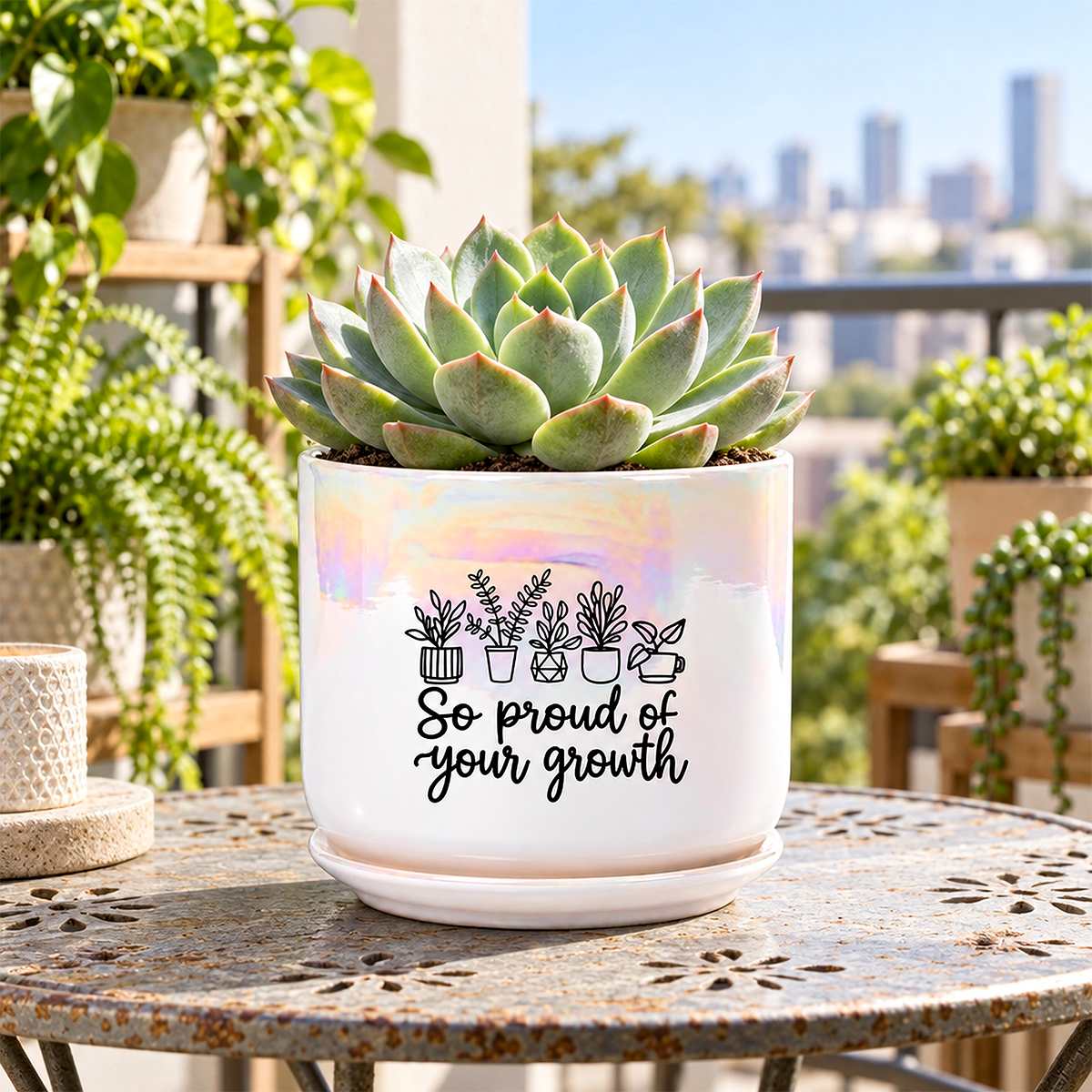 A white ceramic succulent planter with a black printed graphic of potted plants and the text 'So proud of your growth' sits on a metal table in a garden setting with a city skyline in the background.