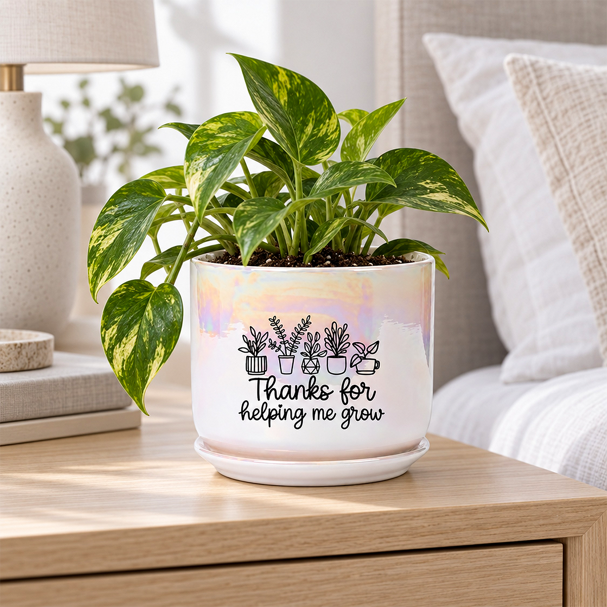 A potted pothos plant in a white ceramic pot with a matching saucer, placed on a wooden bedside table in a bedroom. The pot has a subtle iridescent sheen and is printed with a black line drawing of houseplants and the text 'Thanks for helping me grow'.