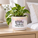 A white ceramic planter pot with a subtle iridescent sheen, featuring a black graphic of potted plants and the text 'Thanks for helping me grow' on the front. The pot sits on a dark wooden table in a bright, modern living room setting with a window, plants, and curtains in the background.