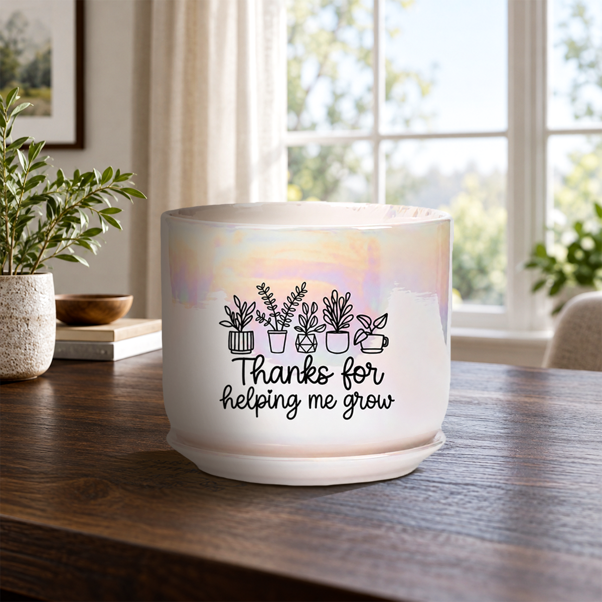 A white ceramic planter pot with a black graphic of potted plants and the text 'Thanks for helping me grow' sitting on a wooden table in a bright, modern room.