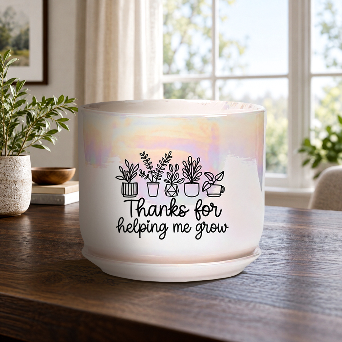 A white ceramic plant pot with a subtle iridescent sheen, featuring a black line art graphic of potted plants and the text 'Thanks for helping me grow' on the front. The pot sits on a dark wooden table in a bright, modern room with a window, plants, and curtains in the background.