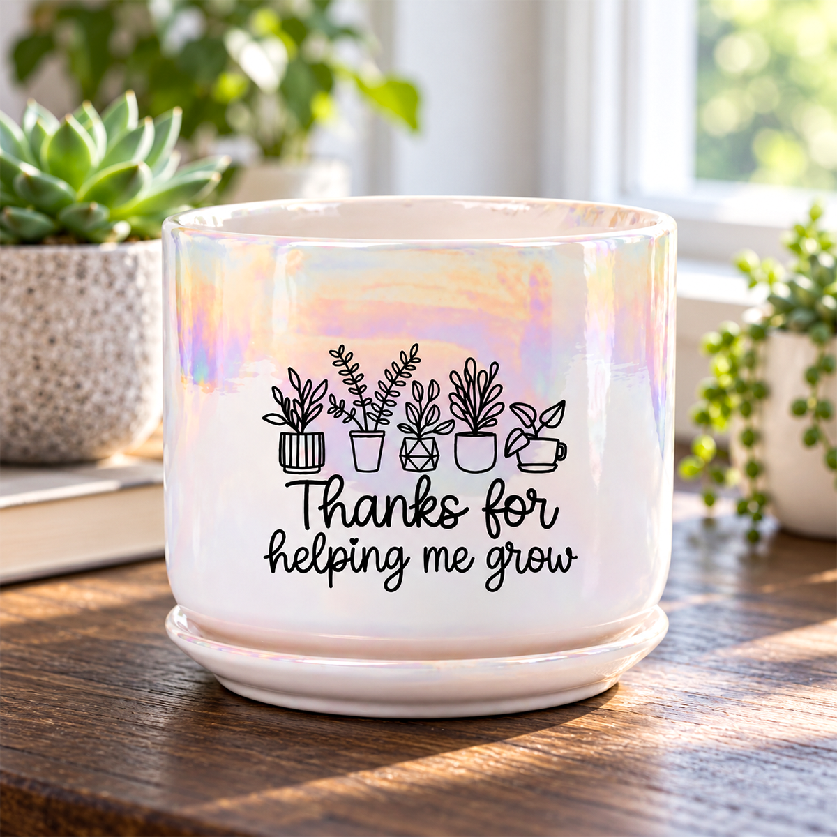 A white ceramic planter pot with a matching saucer, featuring an iridescent finish and a black graphic of potted plants with the text 'Thanks for helping me grow.' It sits on a wooden table in a sunlit room with plants in the background.