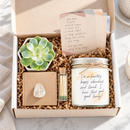 A gift box set containing a jar candle with a handwritten label, a small succulent in a white pot, and a matchstick tube, all nestled in brown crinkle paper inside an open white cardboard box.