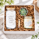 A gift box set containing a jar candle with a handwritten label, a small succulent in a white pot, and a matchstick tube, all nestled in brown crinkle paper inside an open white cardboard box.