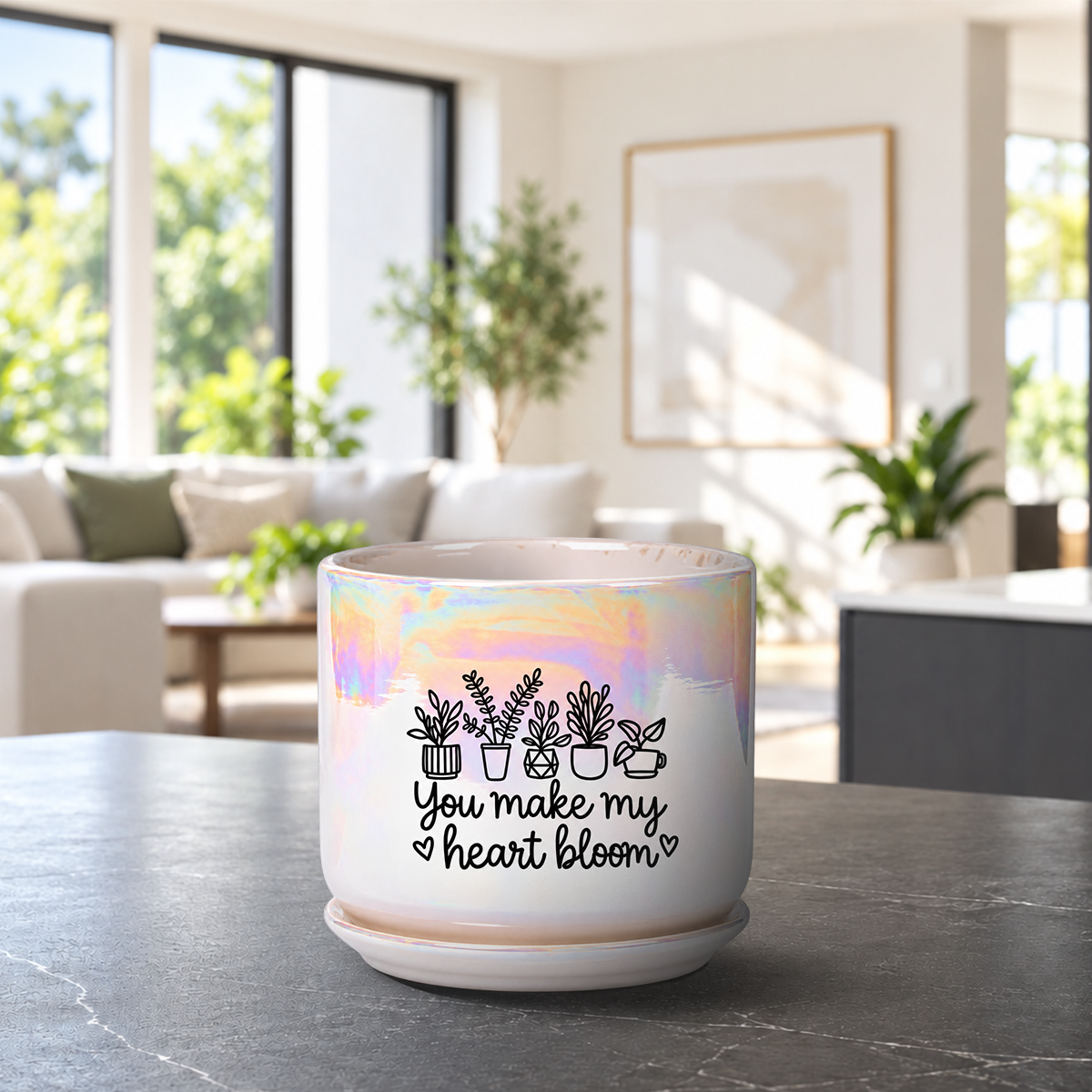 A ceramic planter pot with a matching saucer sits on a dark countertop in a bright, modern living room. The pot is white with a holographic iridescent finish on the upper half, decorated with black line drawings of potted plants and the text 'You make my heart bloom'.