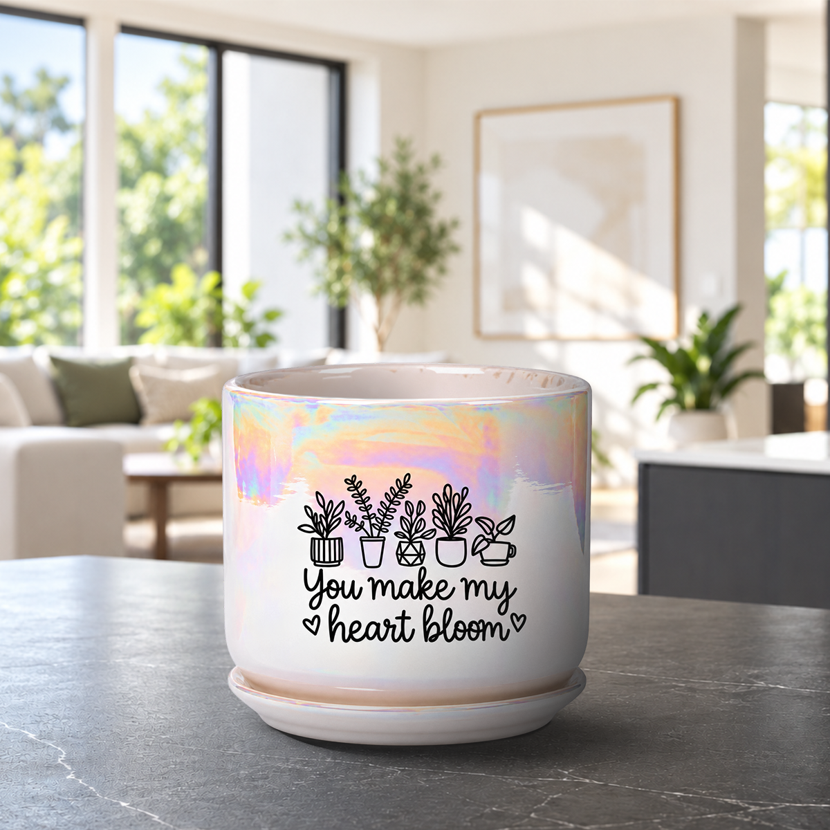 A white ceramic planter pot with a holographic iridescent sheen, featuring a black graphic of potted plants and the text 'You make my heart bloom' on the front. The pot sits on a matching saucer on a dark gray marble countertop, with a blurred modern living room in the background.