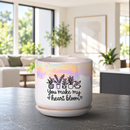 A ceramic planter pot with a matching saucer sits on a dark countertop in a bright, modern living room. The pot is white with a holographic iridescent finish on the upper half, decorated with black line drawings of potted plants and the text 'You make my heart bloom'.