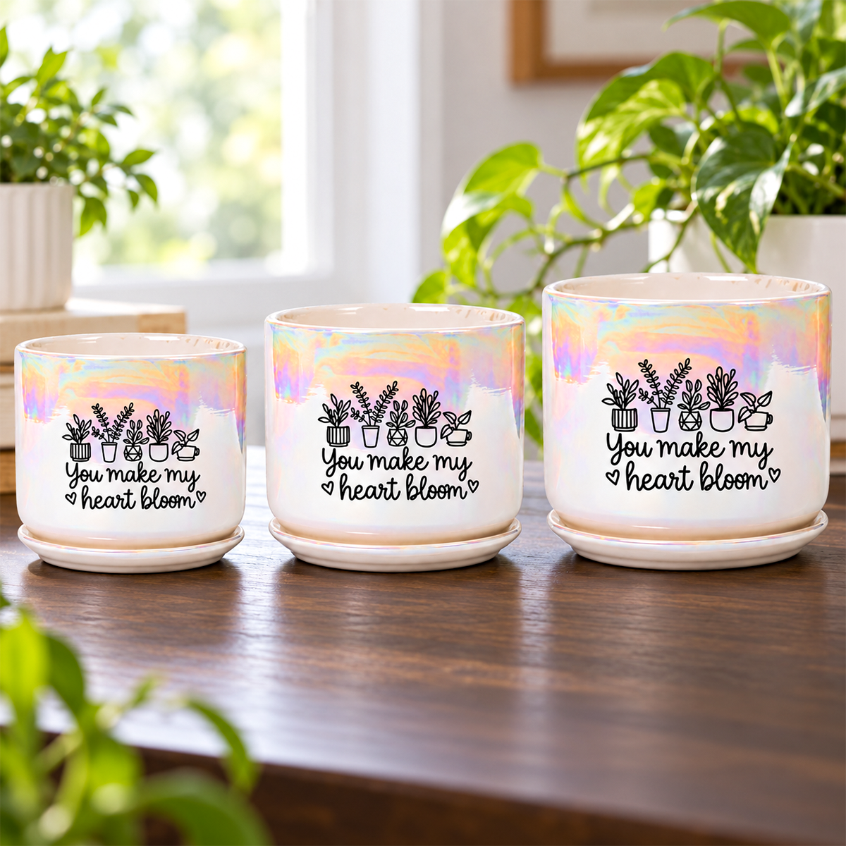 Three small ceramic plant pots with saucers arranged on a wooden table. The pots have an iridescent pastel finish and are printed with black illustrations of potted plants and the phrase 'You make my heart bloom' on the front.